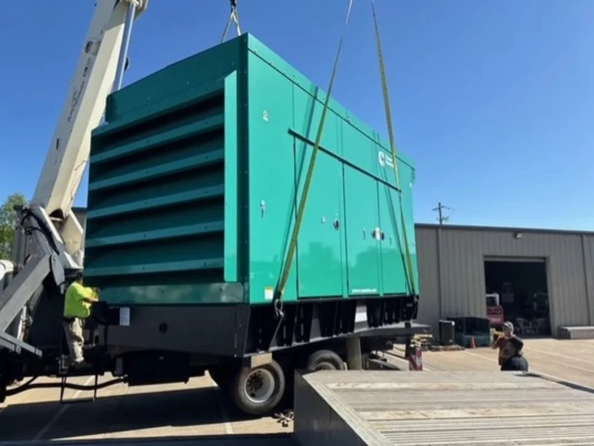 Large green industrial generator being lifted by crane onto a flatbed trailer.