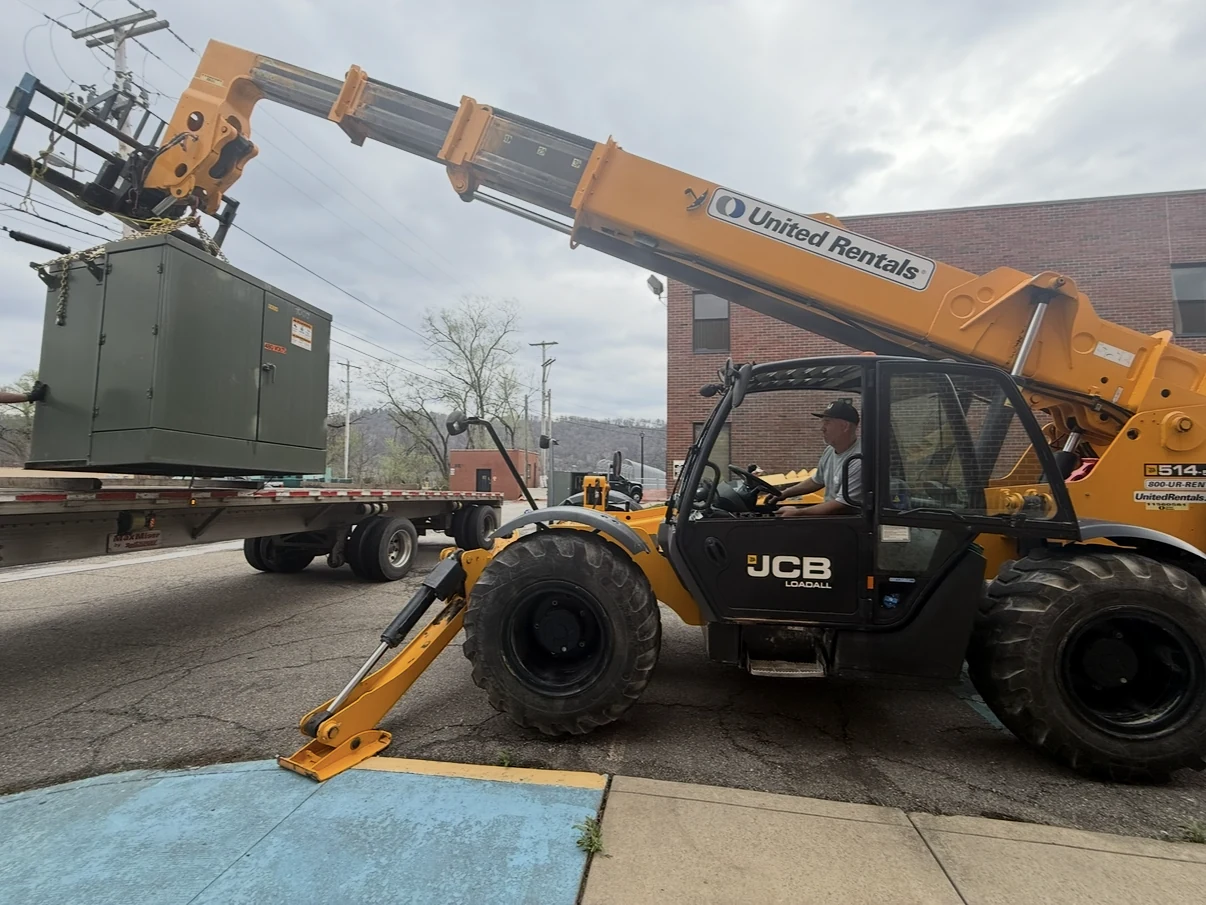 Telehandler lifting a green pad-mounted transformer onto a flatbed.