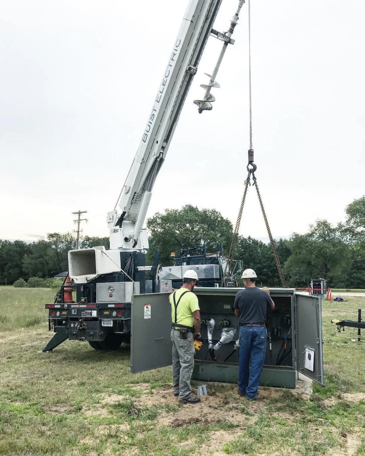 Workers inspecting an open pad-mounted cabinet beside a crane truck.