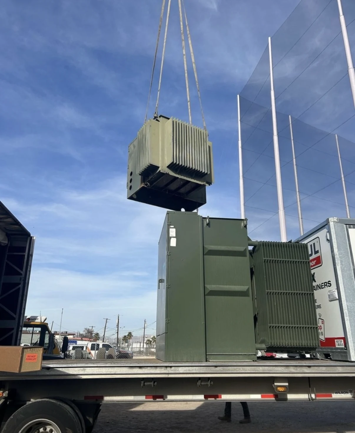 Green industrial transformer suspended above a flatbed by crane rigging.